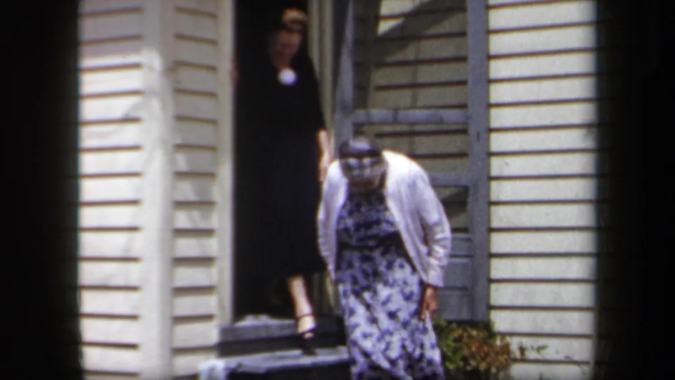 1958 New York home-movie frame showing a woman in a dark dress behind an elderly woman in a floral dress outside a yellow house.
