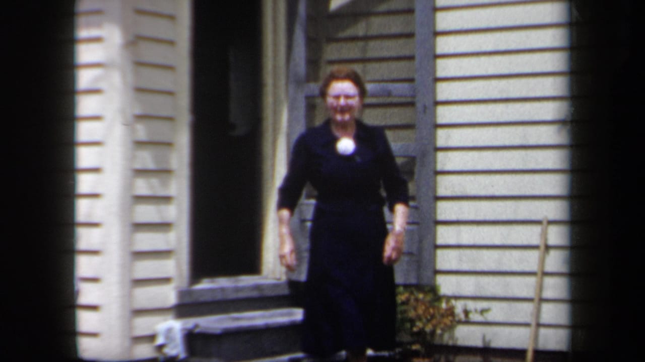 1958 New York home-movie frame of a woman in a dark dress stepping from a light-sided suburban house exterior.