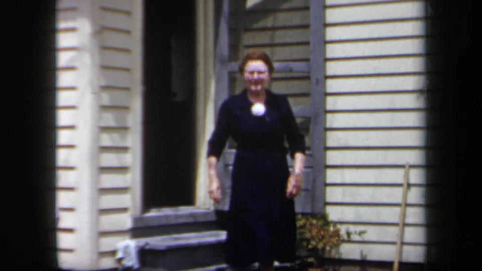 1958 New York home-movie frame of a woman in a dark dress stepping from a light-sided suburban house exterior.