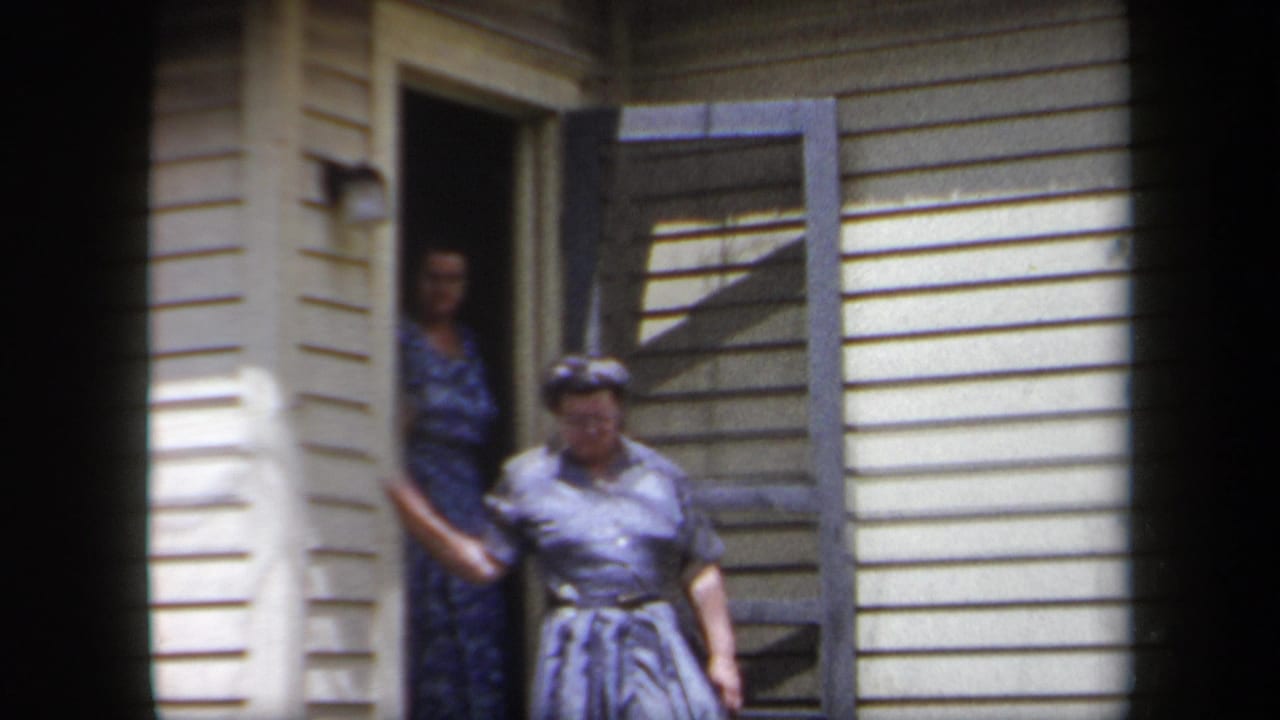 1958 New York home-movie frame showing two adult women at an open porch doorway of a suburban house.