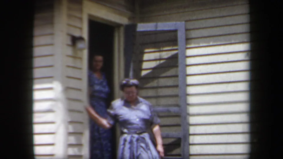 1958 New York home-movie frame showing two adult women at an open porch doorway of a suburban house.