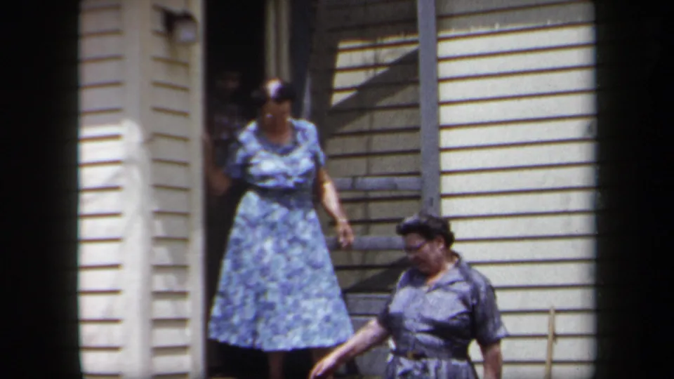 1958 New York home-movie frame with two women outside a suburban house, one exiting the doorway and one walking past.