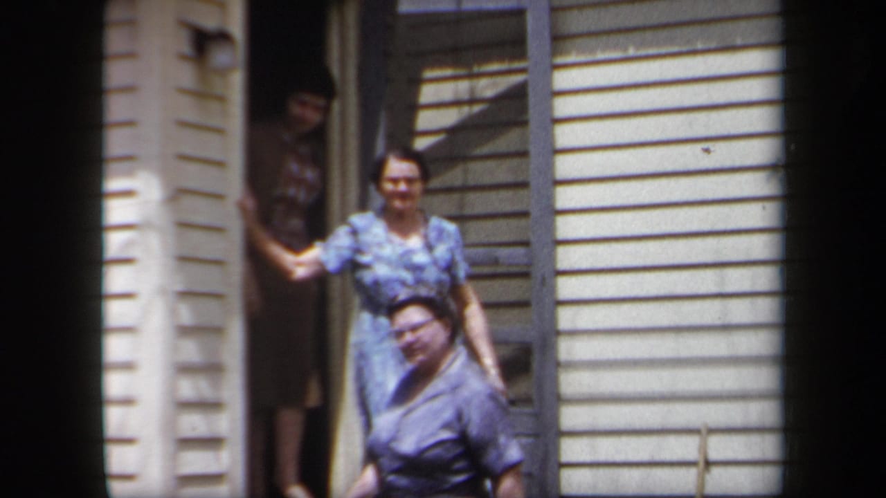 In 1958 New York home-movie footage, women in period dresses gather at a house doorway in bright sunlight.