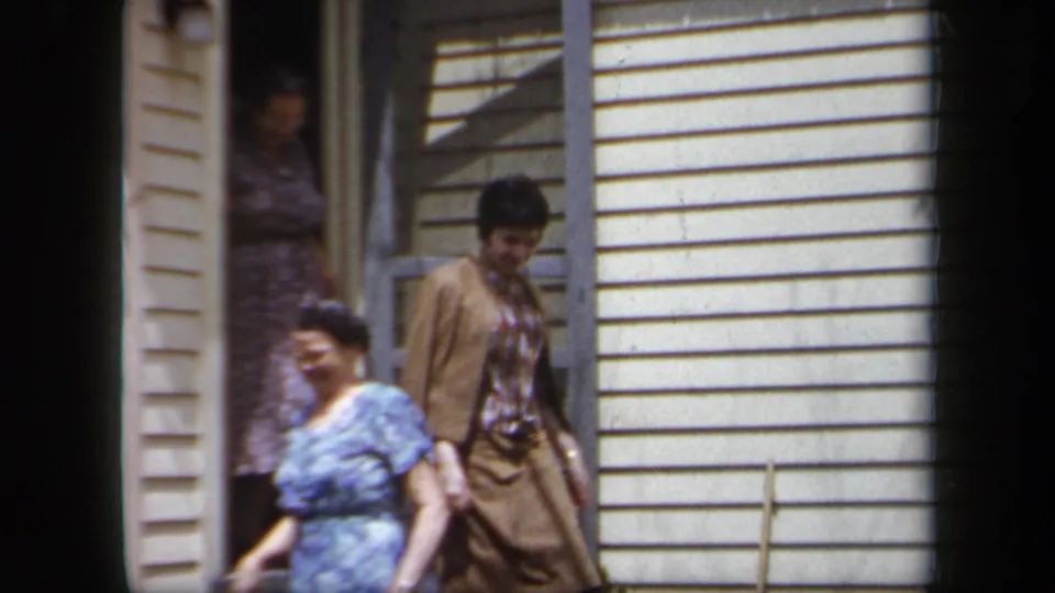 Two women in 1950s clothing move past the doorway of a light-sided New York house in bright daylight.