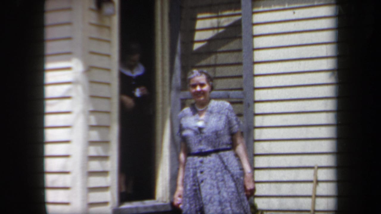 In 1958 New York home-movie footage, an older woman in a blue patterned dress stands near a doorway with another woman partly visible inside.