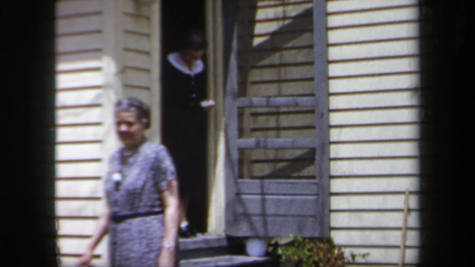 An older woman in a patterned dress walks beside cream siding while a second woman in a dark dress remains in the doorway.