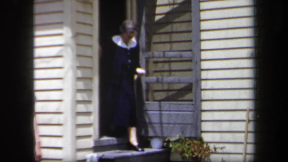 An older woman in a dark dress with a white collar steps from the doorway of a cream-sided house in 1958 New York home-movie footage.