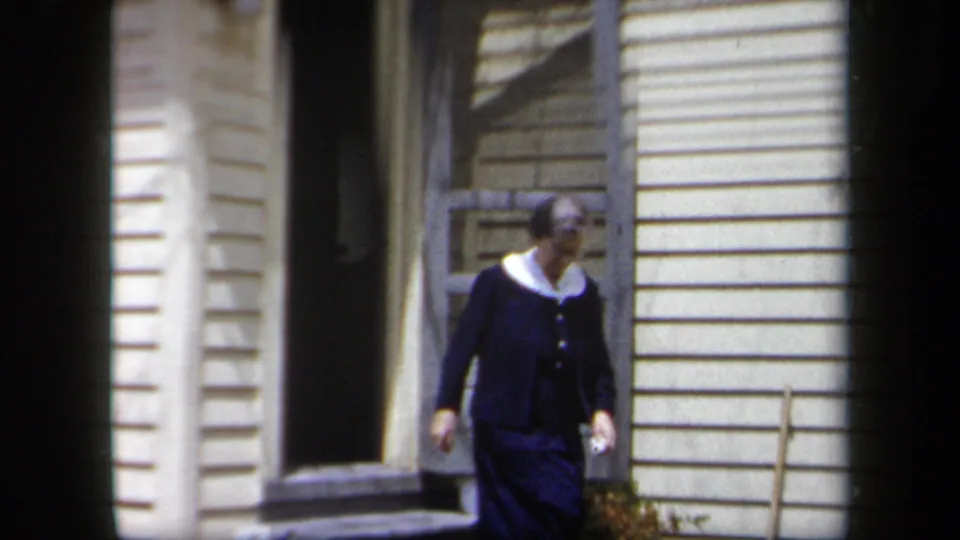 An older woman in a dark dress and white collar walks past the doorway and shuttered window of a cream-sided house.
