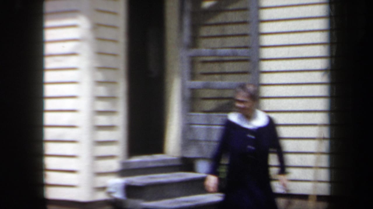 1958 New York home-movie frame showing a woman near porch steps outside a clapboard house with soft focus and handheld framing.