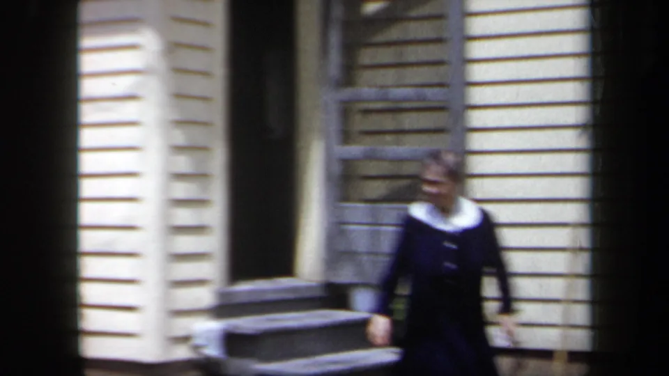 1958 New York home-movie frame showing a woman near porch steps outside a clapboard house with soft focus and handheld framing.