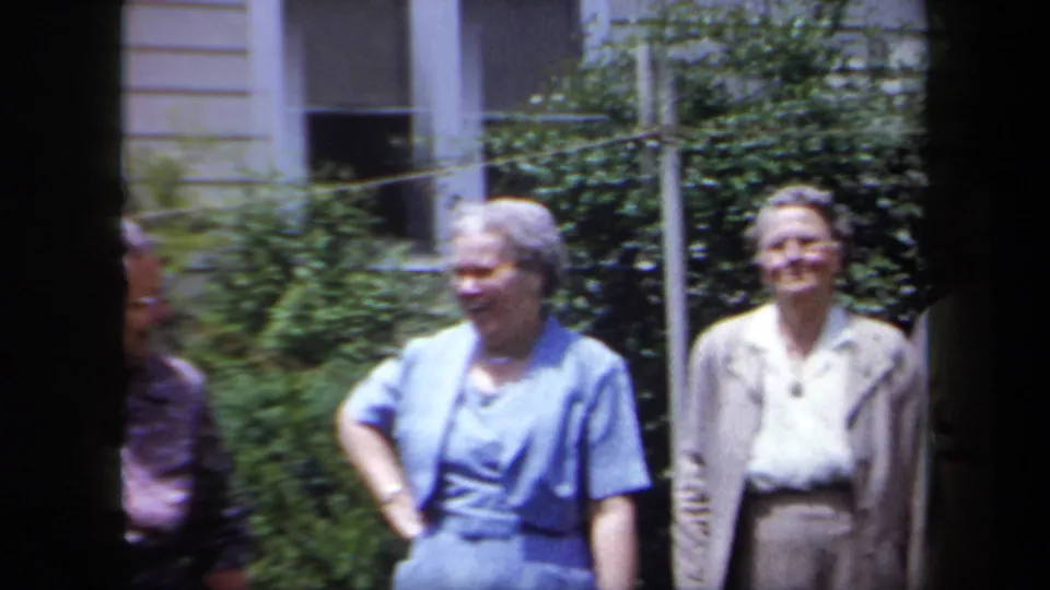 1958 New York home-movie frame of women standing together by garden shrubs outside a residential house.