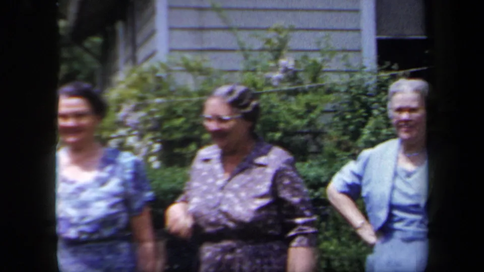 Older women stand and smile outside a New York house in 1958, framed by greenery and clapboard siding.