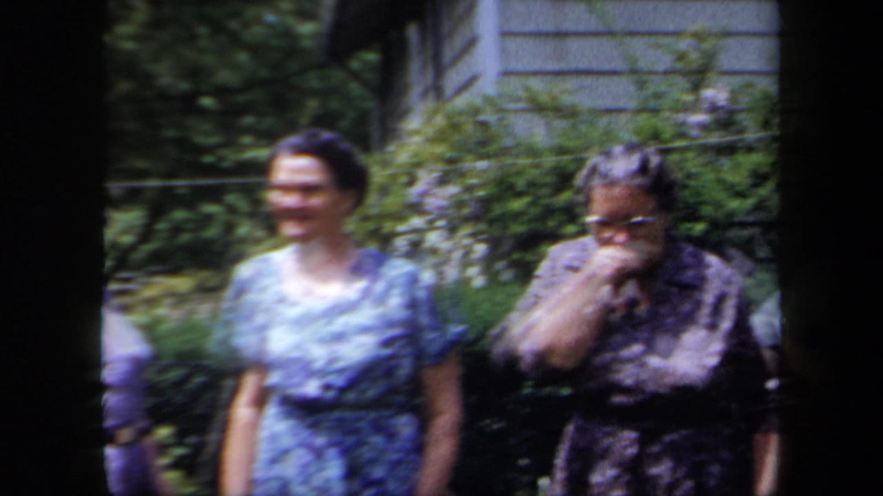 1958 New York home-movie frame showing older women in patterned dresses outside a suburban house in bright sun.