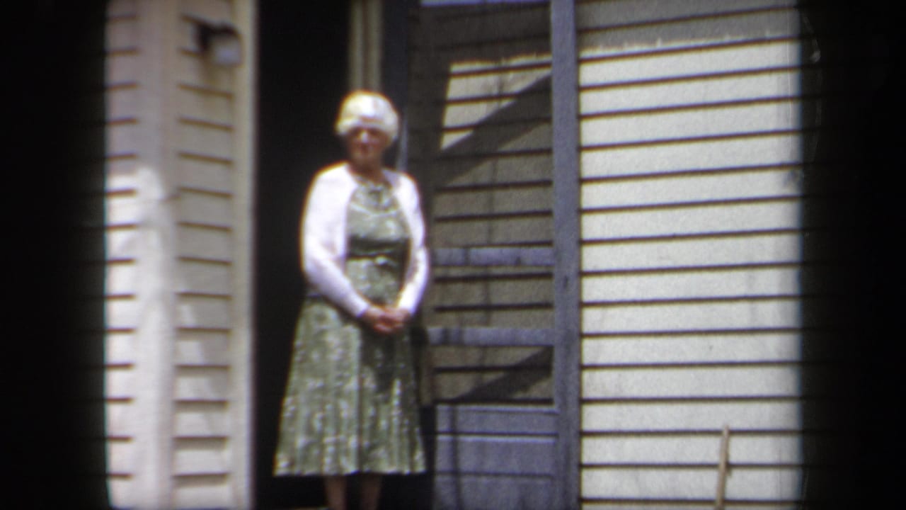 1958 New York home-movie frame of an older woman in a green dress and white cardigan standing at a residential doorway.