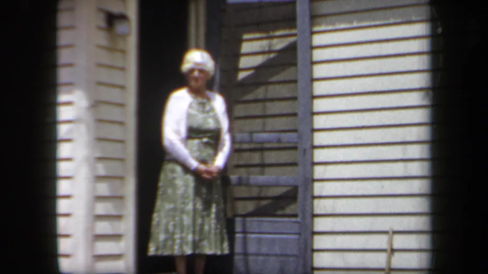 1958 New York home-movie frame of an older woman in a green dress and white cardigan standing at a residential doorway.
