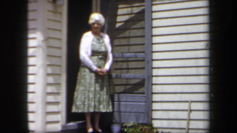1958 New York home-movie frame showing an older woman waiting at the threshold of a suburban home.
