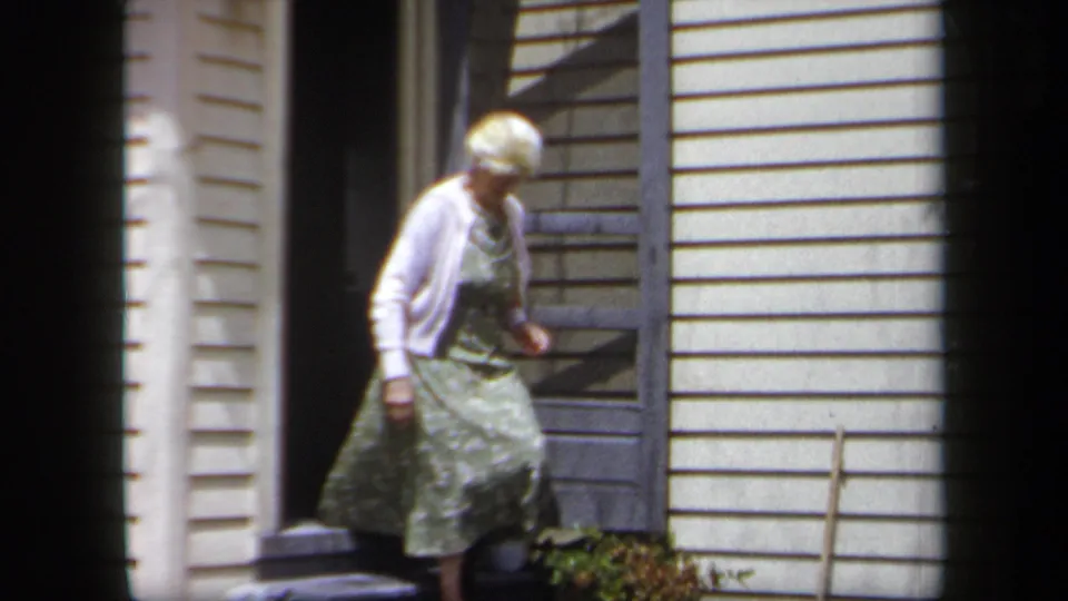 1958 New York home-movie frame of an older woman stepping from a residential doorway in bright daylight.