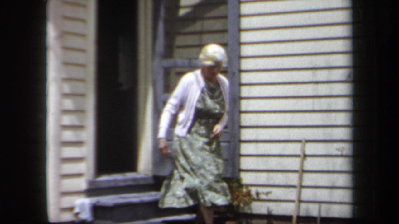 In 1958 New York home-movie footage, an older woman leaves a house with white siding and steps down from the porch.