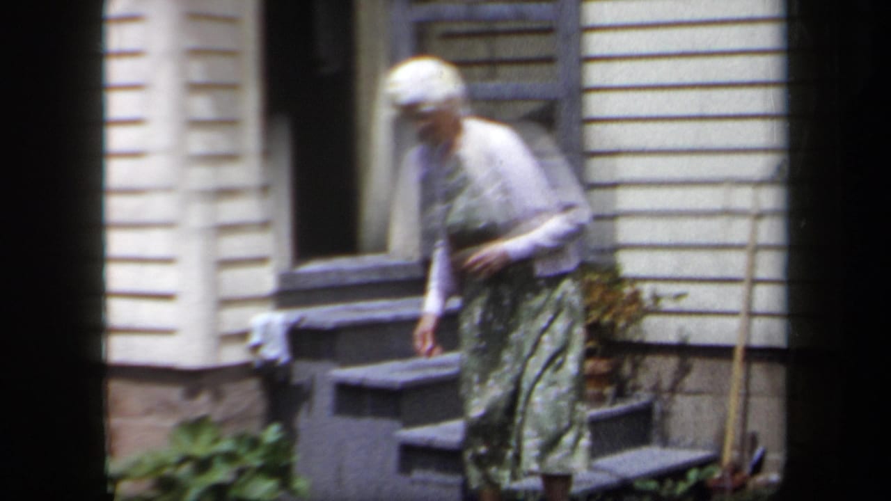 The woman continues down the steps outside the white-sided house, filmed with soft 8mm color and slight blur.