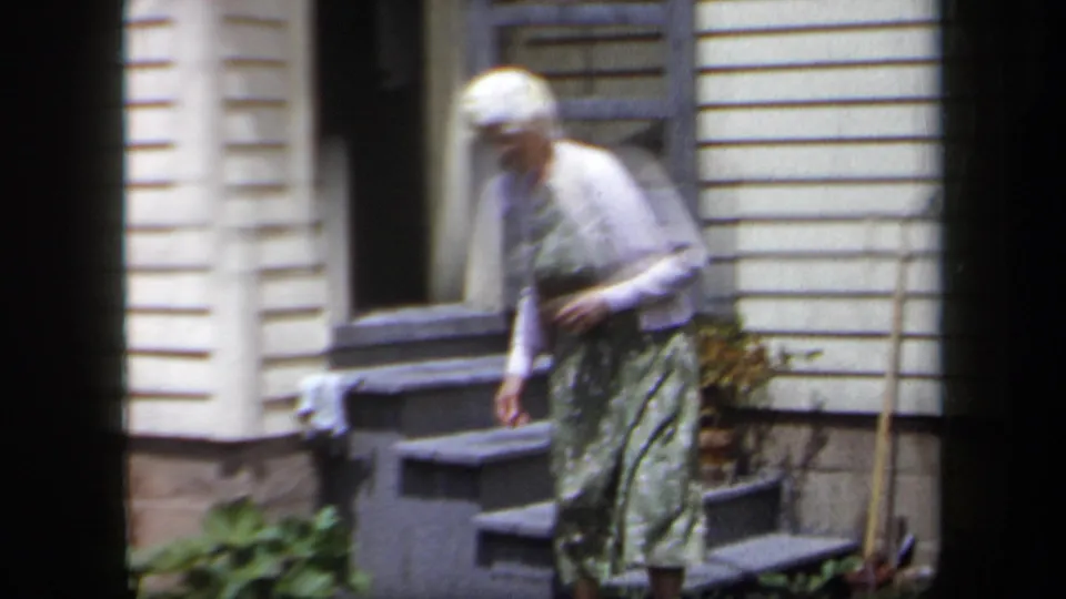 The woman continues down the steps outside the white-sided house, filmed with soft 8mm color and slight blur.