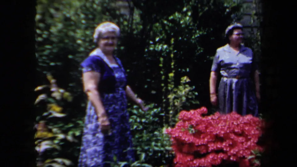 Two adult women stand in a leafy garden with red flowers, facing the camera in a casual home-movie pose.