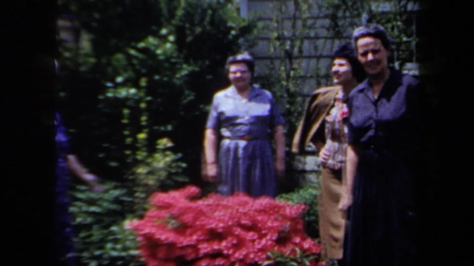 A small group of adults stands together in the garden, with red flowers and dense greenery in the foreground.