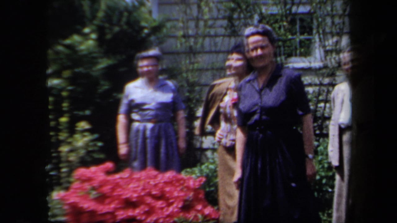 1958 New York home-movie frame showing older women in dresses standing outdoors by flowers, shrubs, and a stone wall.