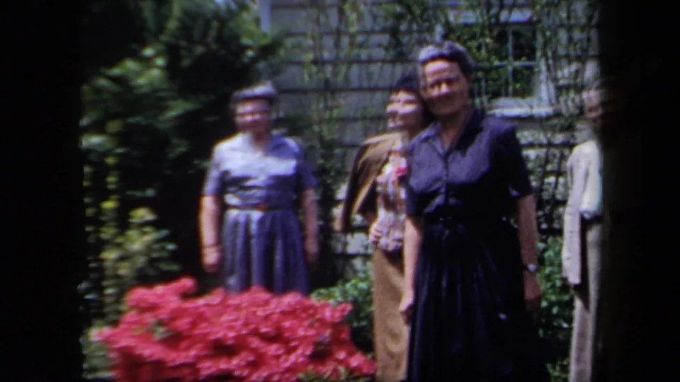 1958 New York home-movie frame showing older women in dresses standing outdoors by flowers, shrubs, and a stone wall.