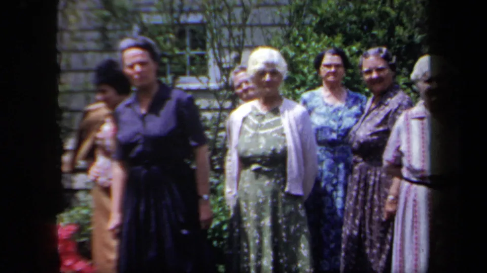 Home-movie frame of older women posing outdoors in dresses, with garden plants, a stone wall, and partial figures at the edges.