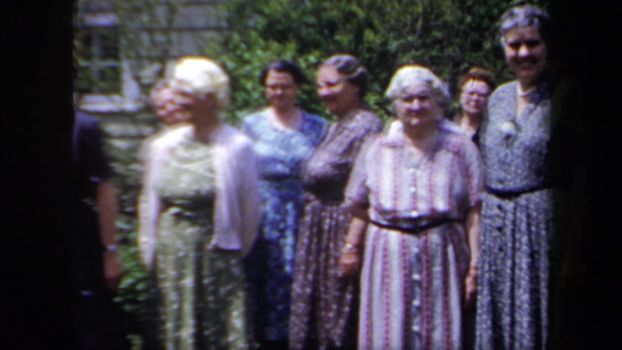 Women in 1950s-style dresses stand in a New York garden, with foliage and part of a house or stone wall behind them.