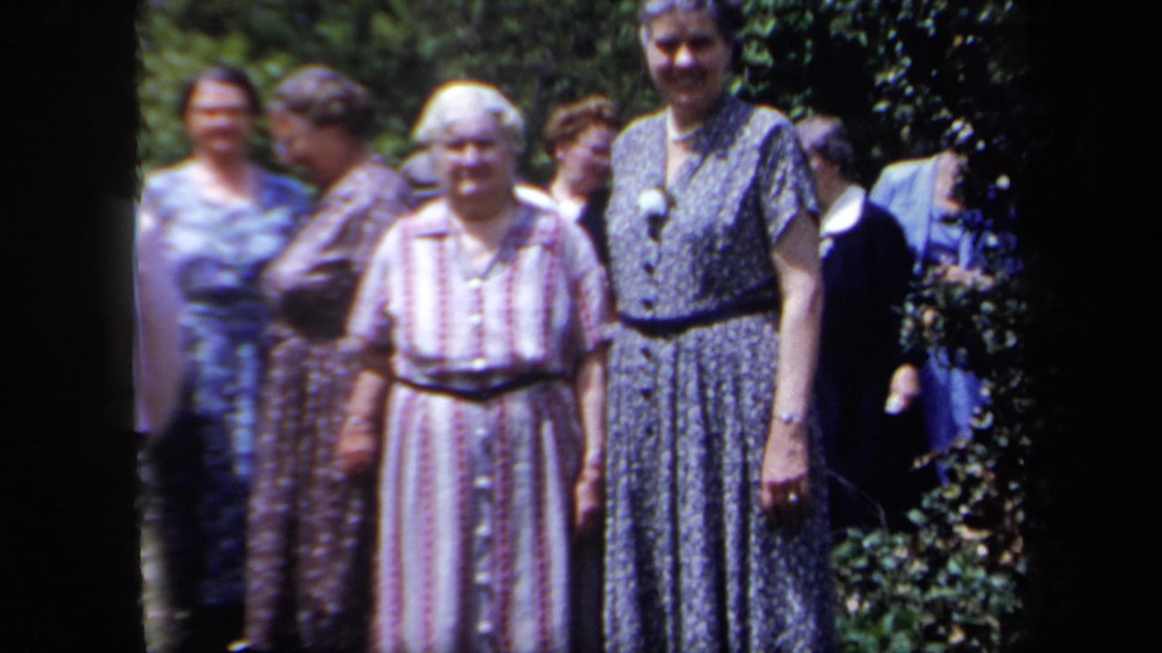 A group of women in vintage dress clothing stands near garden foliage, with faces turned toward the camera.