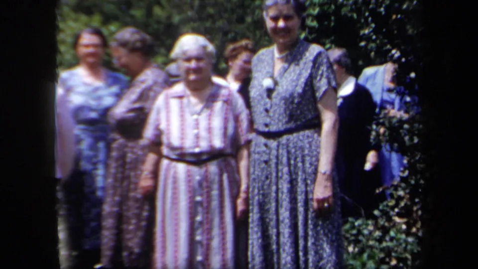 A group of women in vintage dress clothing stands near garden foliage, with faces turned toward the camera.