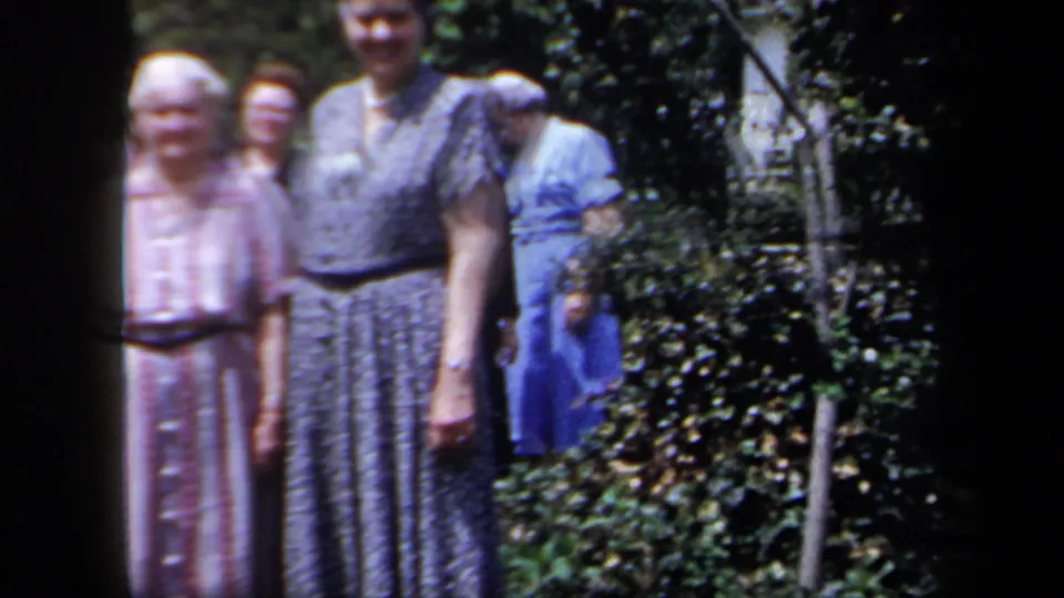 A soft-focus 8mm frame shows women in dress clothing standing in a New York garden, with greenery filling the background.