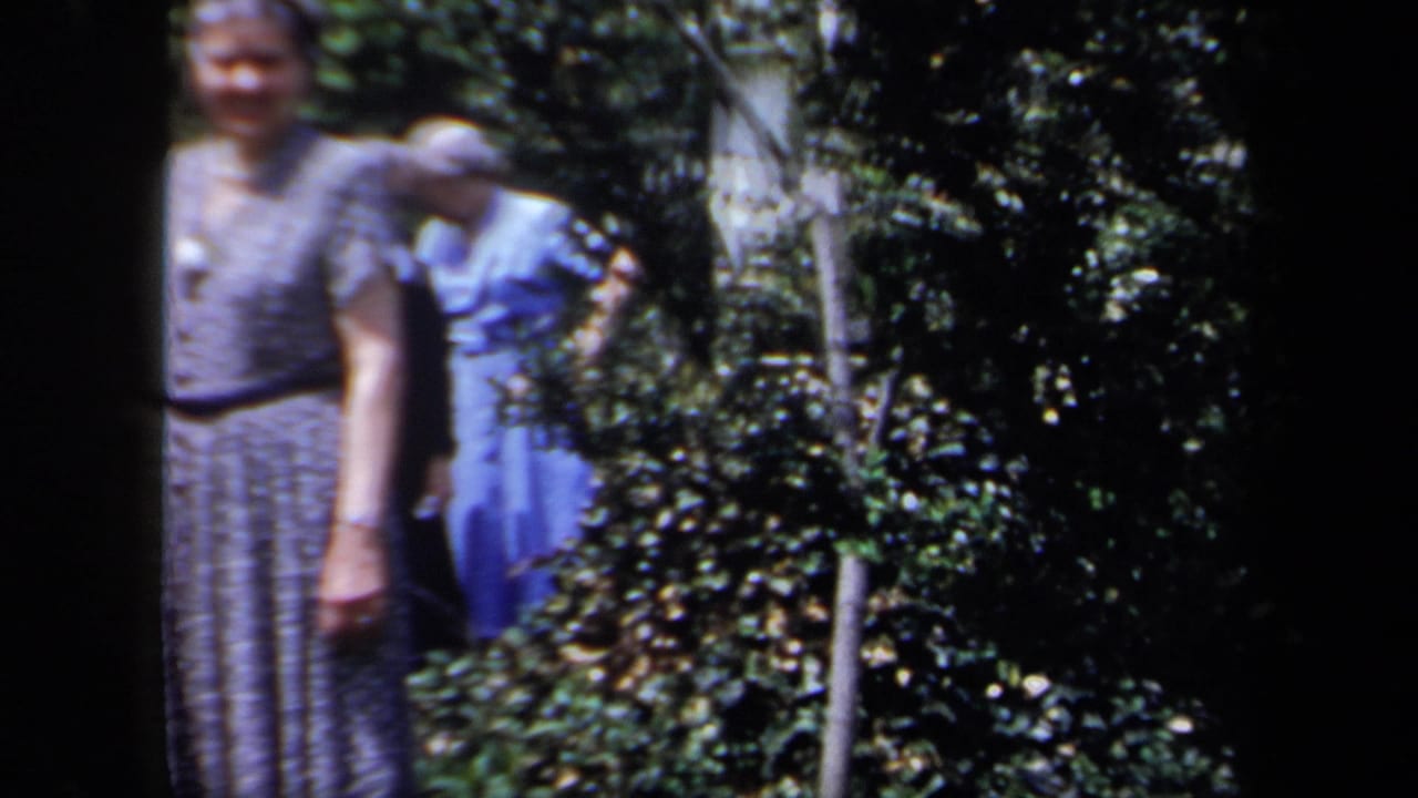 In 1958 New York home-movie footage, two older women in dresses stand near leafy shrubs, with soft focus and dark frame edges.