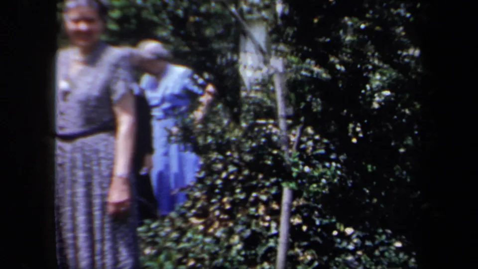 In 1958 New York home-movie footage, two older women in dresses stand near leafy shrubs, with soft focus and dark frame edges.