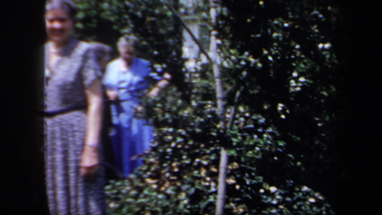 Two older women appear in a leafy garden in 1958 New York footage, with one in blue partly behind shrubs and dark film borders visible.