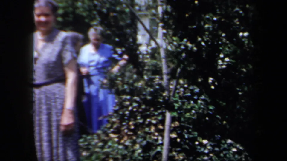 Two older women appear in a leafy garden in 1958 New York footage, with one in blue partly behind shrubs and dark film borders visible.