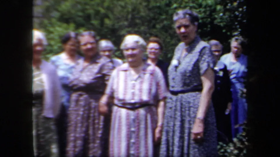 Several older women stand together in a New York garden in 1958 home-movie footage, wearing patterned dresses and facing the camera.