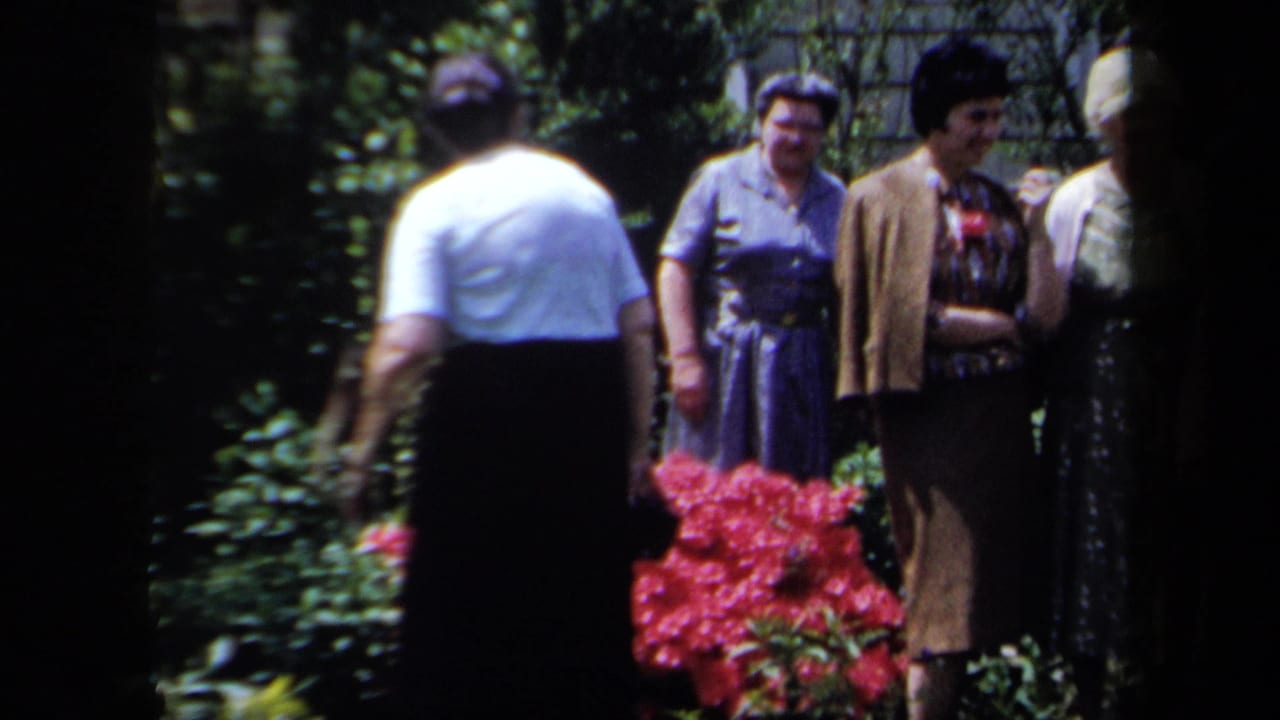Women converse in a garden while one person stands with their back to the camera.