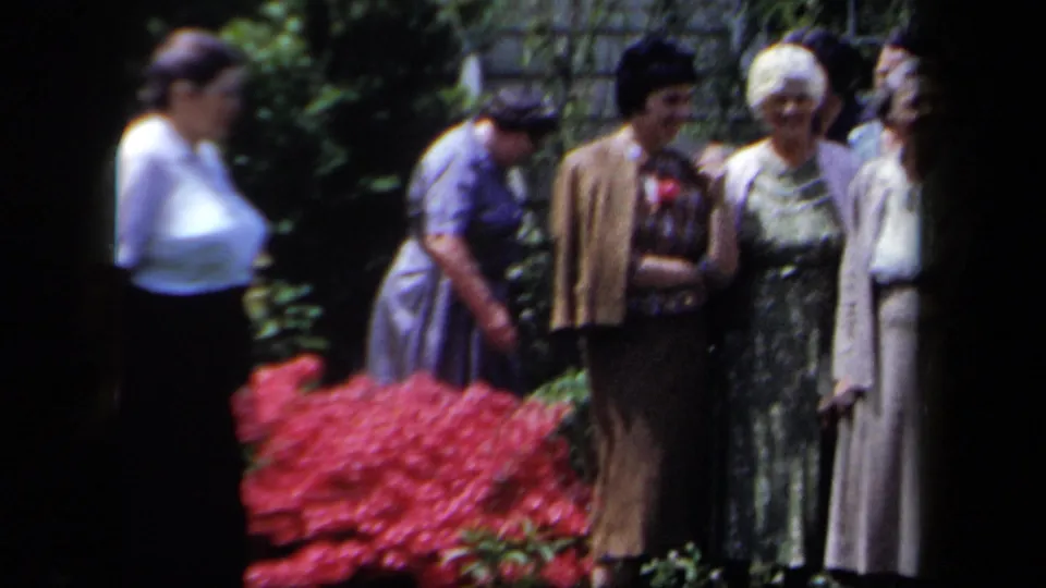 Older women in mid-century clothing stand close together in a New York garden, framed by red flowers and foliage.