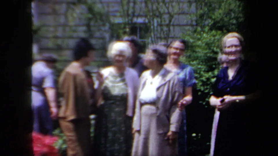 A group of older women in 1950s clothing stand together in a New York garden, with leafy shrubs and a wall in the background.