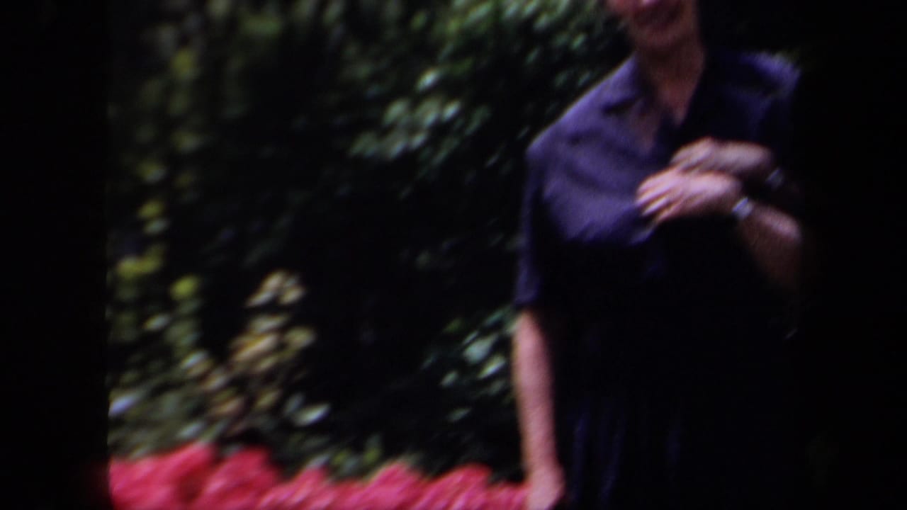 A woman in a dark collared shirt appears at the edge of a New York garden frame with red flowers and leafy shrubs.