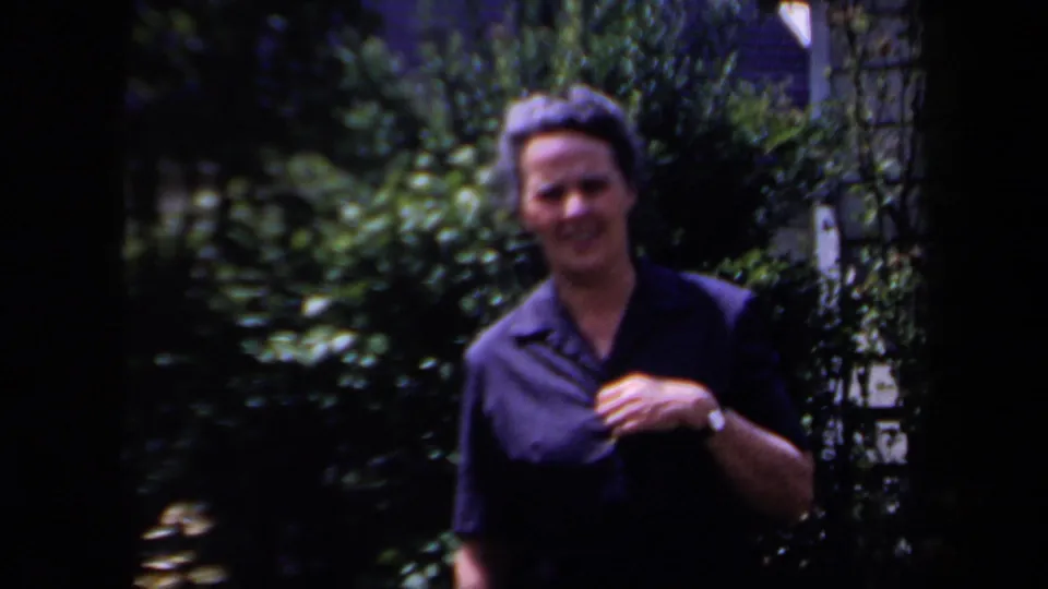 A smiling woman in a dark collared shirt stands in a lush New York garden, seen in soft 1958 8mm home-movie color.