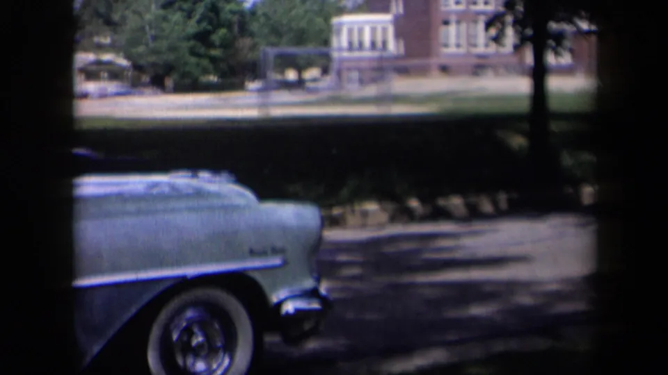 A light blue 1950s car is parked in shade near a street, with a brick building and playground structures beyond.