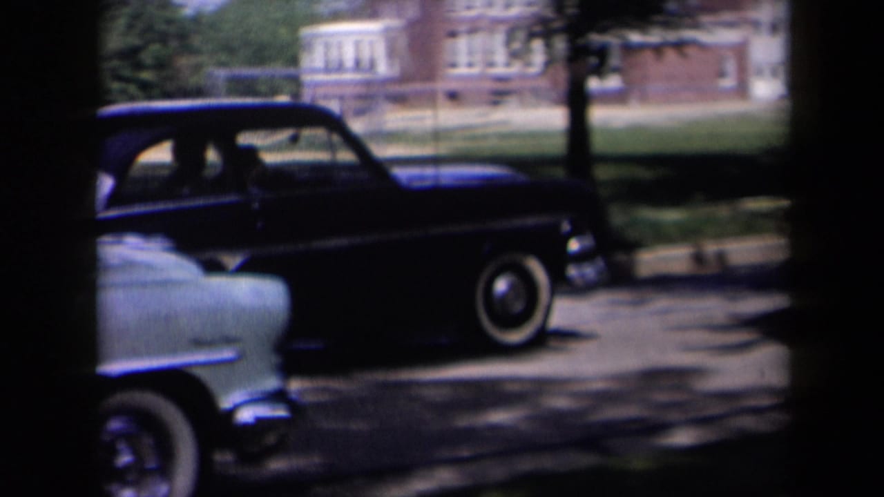 1958 New York street view with a dark station wagon, a light sedan edge, trees, and a brick building in the background.