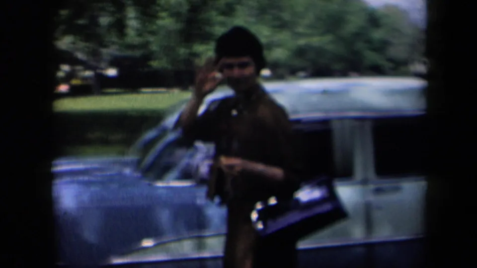 1958 New York home-movie frame of a woman waving beside a light blue sedan, with trees and parked cars behind her.