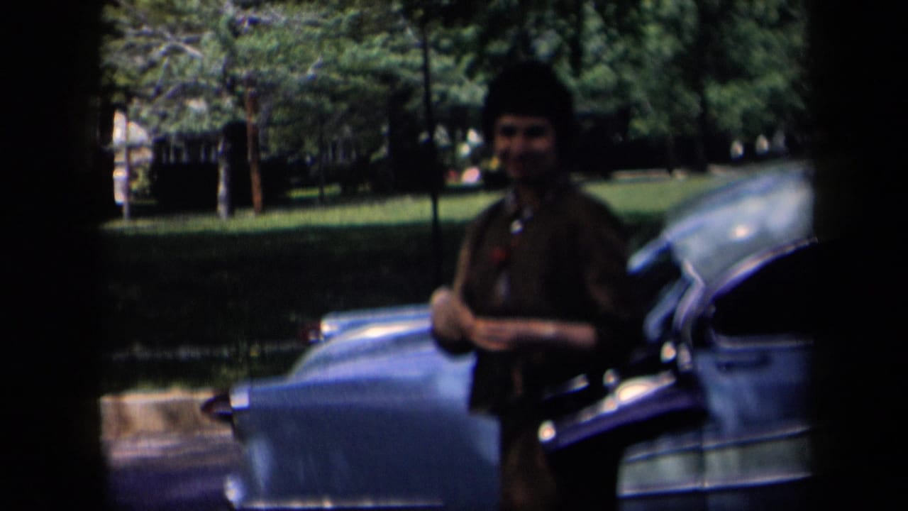 In 1958 New York home-movie footage, a woman in a dark dress and hat stands near a blue parked car beside a green suburban street.