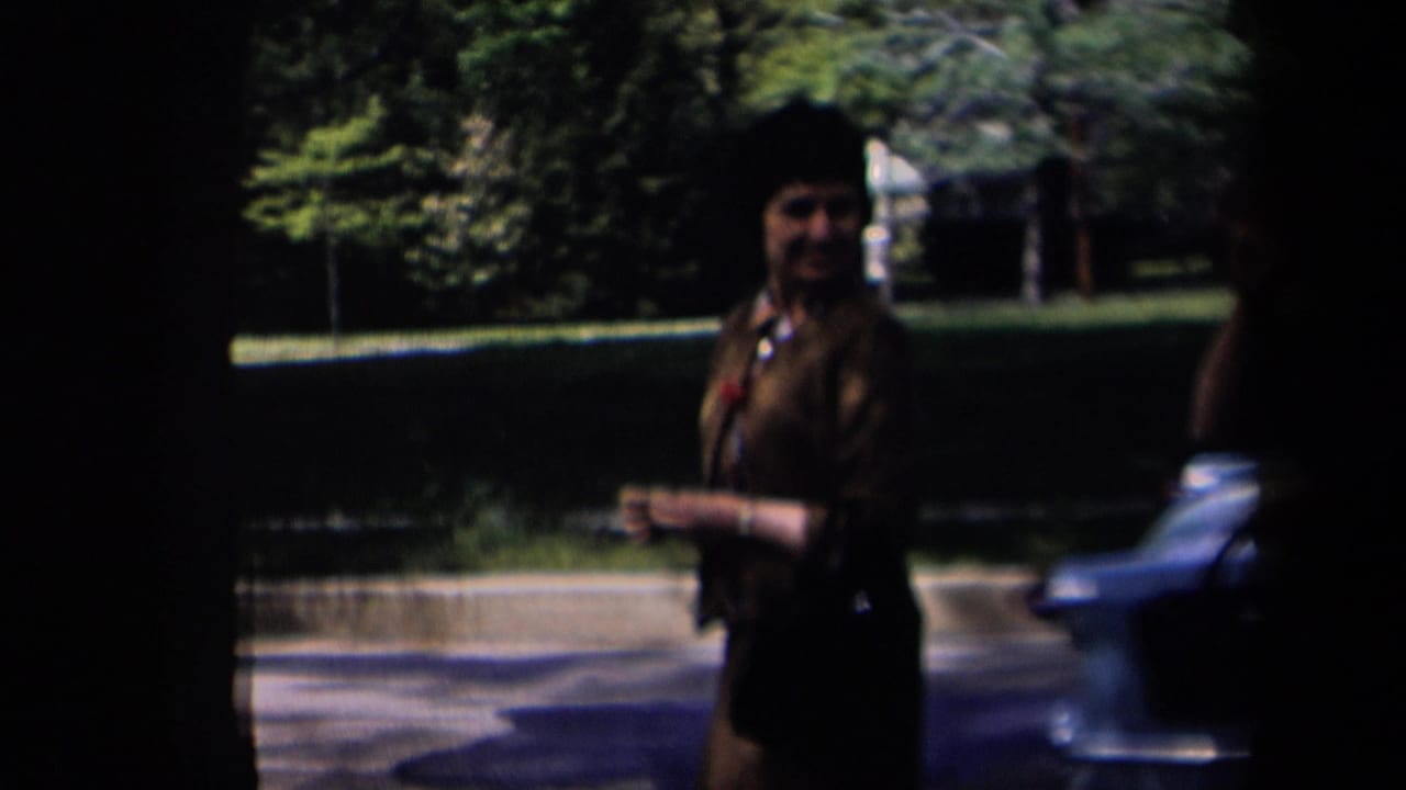The woman walks through a shaded suburban foreground in New York, 1958, with a blue car and green lawn behind her.