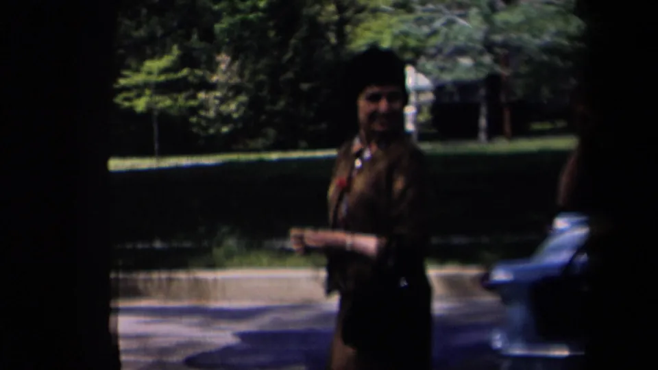 The woman walks through a shaded suburban foreground in New York, 1958, with a blue car and green lawn behind her.