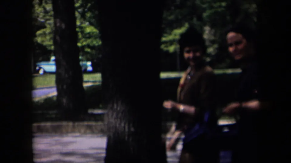 Two adults appear near a tree in a leafy New York residential scene, with a blue car visible across the road in the background.